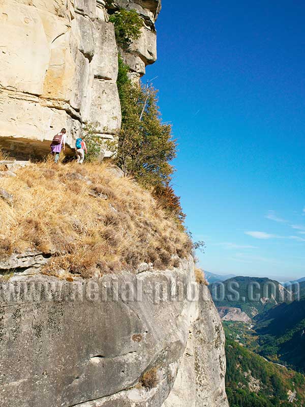 Aerial view of two women hiking on a vertiginous trail and below a rock looking-like a man's face in the Alps. Vue aérienne.